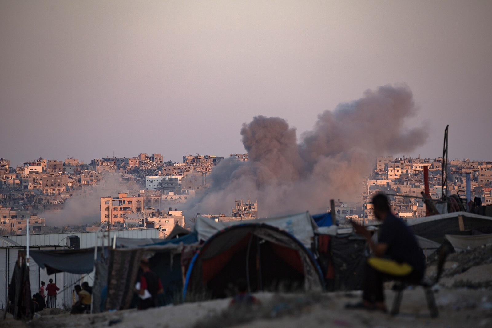 epa11547888 Smoke rises following an Israeli air strike as internally displaced Palestinians sit next to their tents in Khan Younis camp, southern Gaza Strip, 13 August 2024. More than 39,900 Palestinians and over 1,400 Israelis have been killed, according to the Palestinian Health Ministry and the Israel Defense Forces (IDF), since Hamas militants launched an attack against Israel from the Gaza Strip on 07 October 2023, and the Israeli operations in Gaza and the West Bank which followed it.  EPA/HAITHAM IMAD